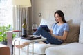 Smiling young woman sitting on sofa with digital tablet and chating with friends, drinking wine.