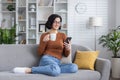 Smiling young woman sitting on sofa at home and resting, using mobile phone and holding cup with drink Royalty Free Stock Photo