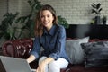 Smiling young woman sitting on sofa with laptop computer and chating with friends