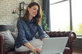 Smiling young woman sitting on sofa with laptop computer and chating with friends