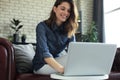 Smiling young woman sitting on sofa with laptop computer and chating with friends