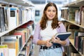 Smiling young woman with stack of books standing in library Royalty Free Stock Photo