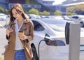Smiling young woman standing on city parking near electric car, charging automobile battery from small city station, drinking coff