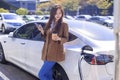 Smiling young woman standing on city parking near electric car, charging automobile battery from small city station, drinking cof