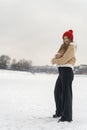 Smiling young woman in stylish beige sheepskin coat with red knitted hat walks in snow outdoors. Winter walking in park