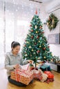 Smiling young woman taking off wrapping paper from giftbox while sitting near christmas tree Royalty Free Stock Photo