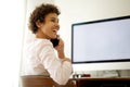 Smiling young woman talking with mobile phone by computer screen Royalty Free Stock Photo