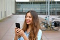 Smiling young woman is using an app in her smartphone device to send a text message while standing in courtyard of office blocks Royalty Free Stock Photo