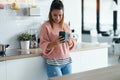 Smiling young woman using her mobile phone while drinking a cup of coffee in the kitchen at home Royalty Free Stock Photo