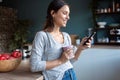 Smiling young woman using her mobile phone while drinking a cup of coffee in the kitchen at home Royalty Free Stock Photo