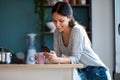 Smiling young woman using her mobile phone while drinking a cup of coffee in the kitchen at home Royalty Free Stock Photo