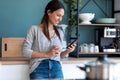 Smiling young woman using her mobile phone while drinking a cup of coffee in the kitchen at home Royalty Free Stock Photo