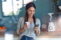 Smiling young woman using her mobile phone while drinking a cup of coffee in the kitchen at home Royalty Free Stock Photo