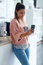 Smiling young woman using her mobile phone while drinking a cup of coffee in the kitchen at home Royalty Free Stock Photo