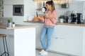 Smiling young woman using her mobile phone while drinking a cup of coffee in the kitchen at home Royalty Free Stock Photo