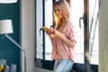 Smiling young woman using her mobile phone while standing next to the window at home Royalty Free Stock Photo