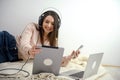 Smiling young woman wearing headphones lying on bed with laptop, teenage student studying online at home, listening to