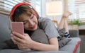 Smiling young woman wearing headphones and using smart phone, resting on couch in cozy living room Royalty Free Stock Photo