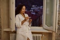 Smiling young woman in white robe, enjoying her morning espresso coffee, standing by opened window at home Royalty Free Stock Photo