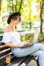 Smiling young woman working on laptop sitting on bench in park and having coffee. Technology, communication, education and remote Royalty Free Stock Photo