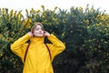 Smiling young woman in a yellow raincoat and hood with closed eyes enjoying the moment on walk with blooming bushes Royalty Free Stock Photo