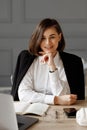 Smilling business lady with glasses on her eyes, wearing a white shirt and a black jacket on her shoulders, is sitting at a desk