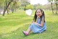 Smilling little Asian girl holding blank heart label lying on green grass at summer garden