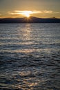 Glassy water with the golden sun setting behind flat mountains in the distance looking from Lakeside Beach at Lake Tahoe