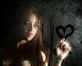 Smooth portrait of model, posing behind transparent glass covered by water drops and draws a heart on the glass Royalty Free Stock Photo