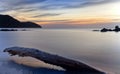 Smooth sea at sunrise, with dramatic moving sky, hills, rocks and tree trunk on beach, port nou, mallorca, spain