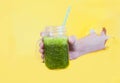 Smoothie from fresh herbs in a glass jar is held by a man`s hand in a hole on a yellow background