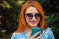 Sms. Young red head Woman in blue jeans texting looking talking to her Mobile Phone While Walking in a City park, green outdoors Royalty Free Stock Photo