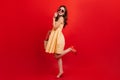 Snapshot full-length of pretty girl in short yellow dress on red background. Woman with dark wavy hair in sunglasses is