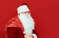 Sneering Santa Claus with bag of gifts stands on red background and looks into camera, closeup portrait. Isolated. Cunning Santa