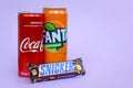 Snickers chocolate bar in brown wrapping lies with red Coca Cola and orange Fanta tin can on lilac pastel background. Famous drink