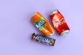 Snickers chocolate bar wrapping lies with empty red Coca Cola and orange Fanta tin cans on lilac pastel background. Famous drink