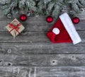 Snow covered evergreen branches plus bright red ornaments, Santa cap and gift box on faded wooden planks for a merry Christmas or Royalty Free Stock Photo