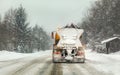 Snow covered orange highway maintenance gritter truck on slippery road, heavy snowing and trees in background