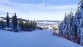 A snow-covered piste for skiers and snowboarders, equipped with snow cannons and safety fences, runs through a spruce forest. Hous