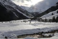 snow covered valley with a farmhouse and a single cross country skiier in the distance while the sun comes through the dark clouds