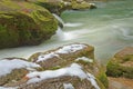 Snow on mossy rocks beside smooth cool flowing river water