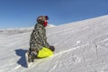Snowboarder in full gear is sitting in the snow after falling against the backdrop of mountains and a blue sky.