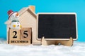 Snowman sitting on wooden block calendar set on 25 December beside blackboard on white wool in front of Wooden house. Copy space f