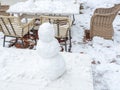 Snowman stands on a wooden table against the background of a snow-covered table and chairs of a street cafe