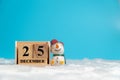 Snowman wearing red hat sitting beside wooden block calendar set on the Christmas date 25 december on white wool and blue backgrou