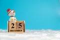 Snowman wearing a red hat sitting on wooden block calendar set on the Christmas date 25 december on white wool and blue background