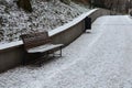 Snowy benches near the supporting concrete gray wall in the park. Paving and metal low fences protect ornamental flower beds from