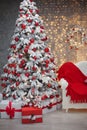 Snowy Christmas tree with red gifts before bokeh lights and wreath decoration on the wall with present boxes in the comfy room in Royalty Free Stock Photo