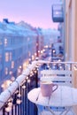 Snowy city balcony at night adorned with glowing string lights, featuring a cozy cup on a table, creating a warm and Royalty Free Stock Photo