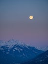 Snowy winter alpine landscape after sunset, pink purple sky with a full moon over a mountain peak